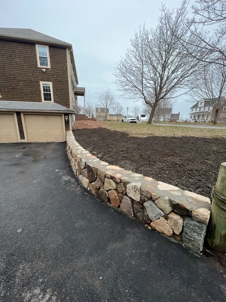 A new stone retaining wall curves along a paved driveway next to a brown shingled house with a yard covered in dark mulch.