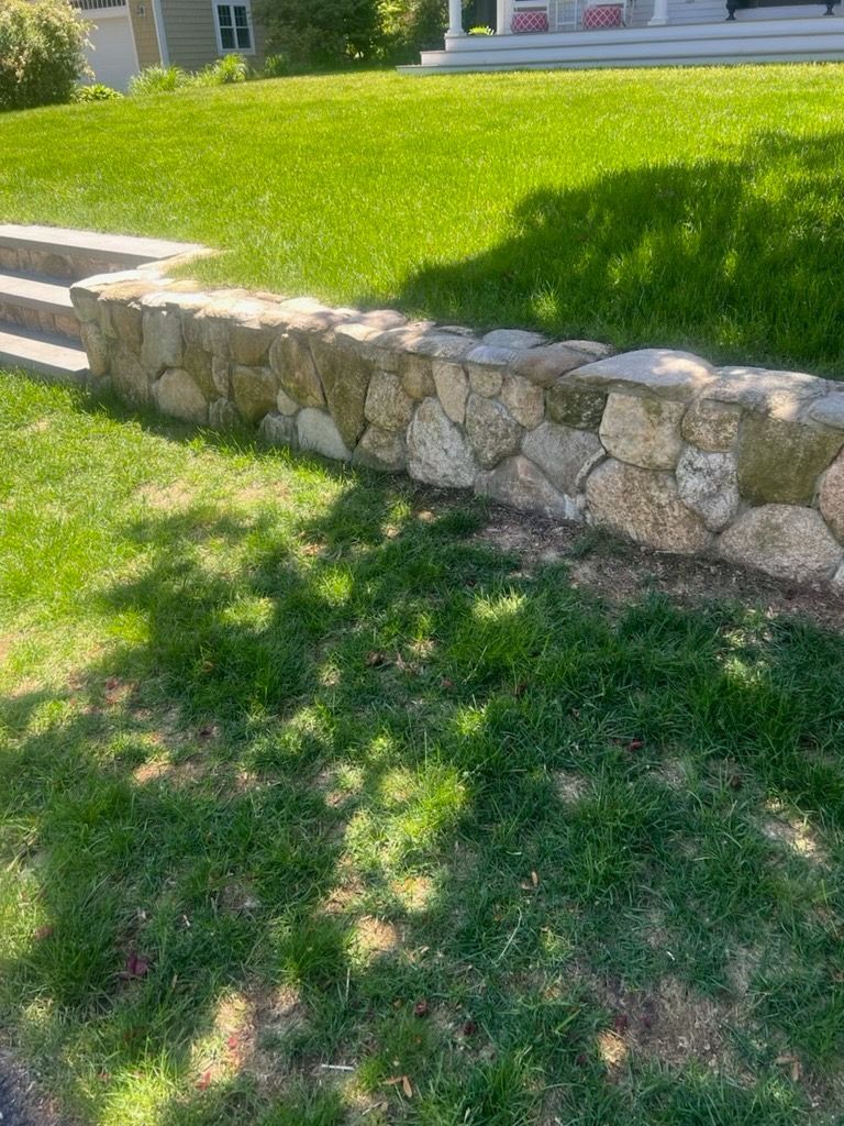 A stone retaining wall built into a grassy hill leads toward a house porch.