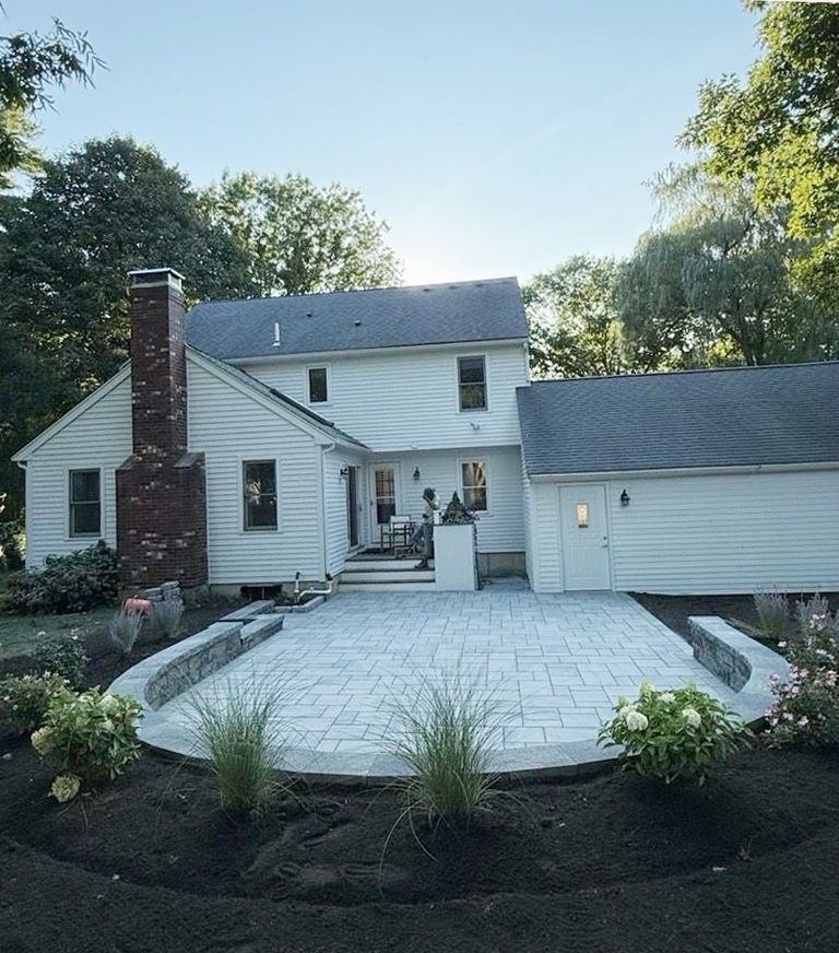 A backyard featuring a stone patio, brick chimney, white siding, and landscaped garden beds with grasses and shrubs.