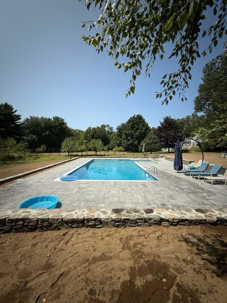 A rectangular swimming pool surrounded by a stone patio on a sunny day, with trees and an unfinished dirt yard nearby.