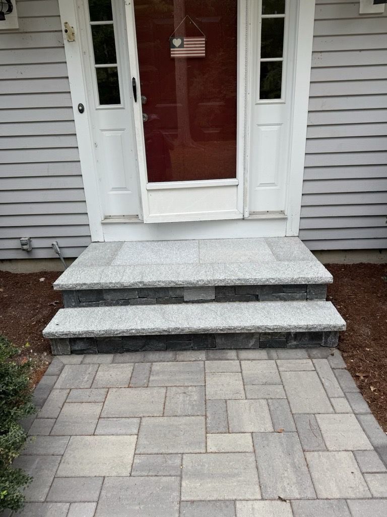 A front door with a red storm door and an American flag sign, accessed by two granite steps and a paved stone walkway.