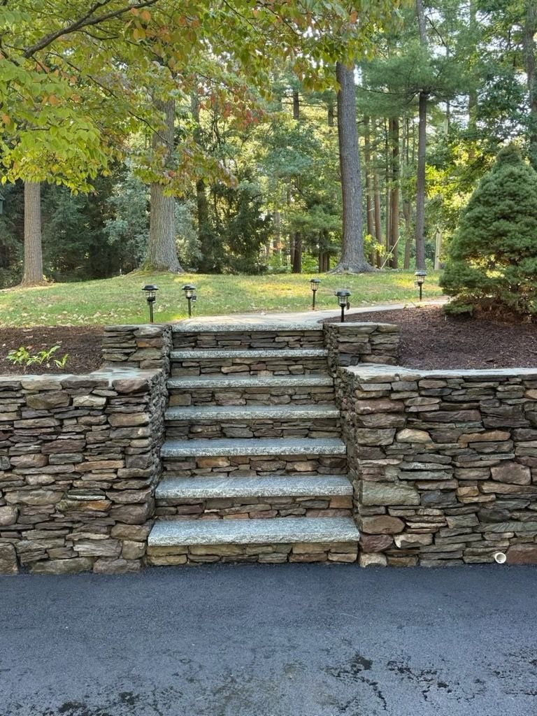 Stone stairs with matching retaining walls leading to a grassy, wooded yard with small landscape lights.
