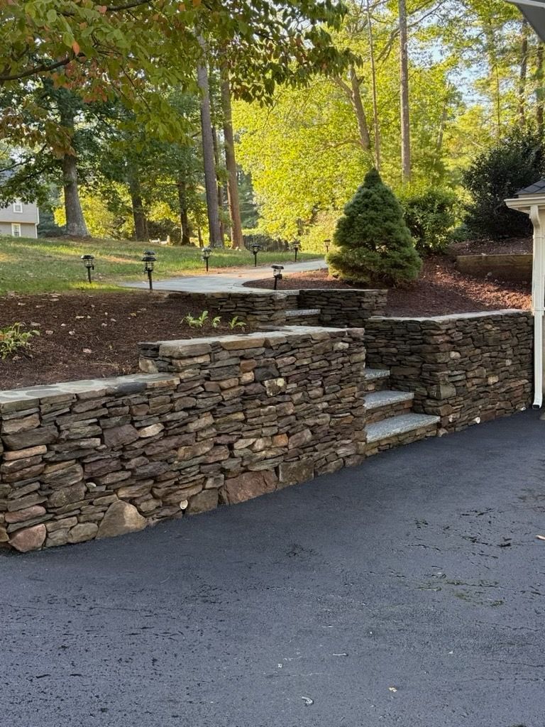 A stone retaining wall with integrated steps lines a driveway in a landscaped yard with trees and mulch.