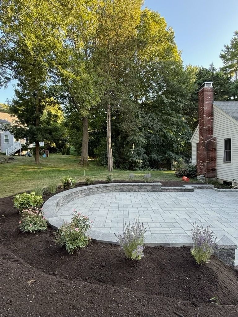 A new light-gray paver patio with a curved retaining wall, surrounded by fresh mulch and young shrubs, next to a house.