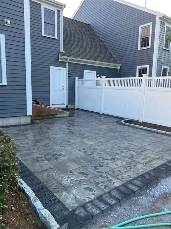 A newly installed grey paver patio with a dark stone border, adjacent to a blue house with a white fence.
