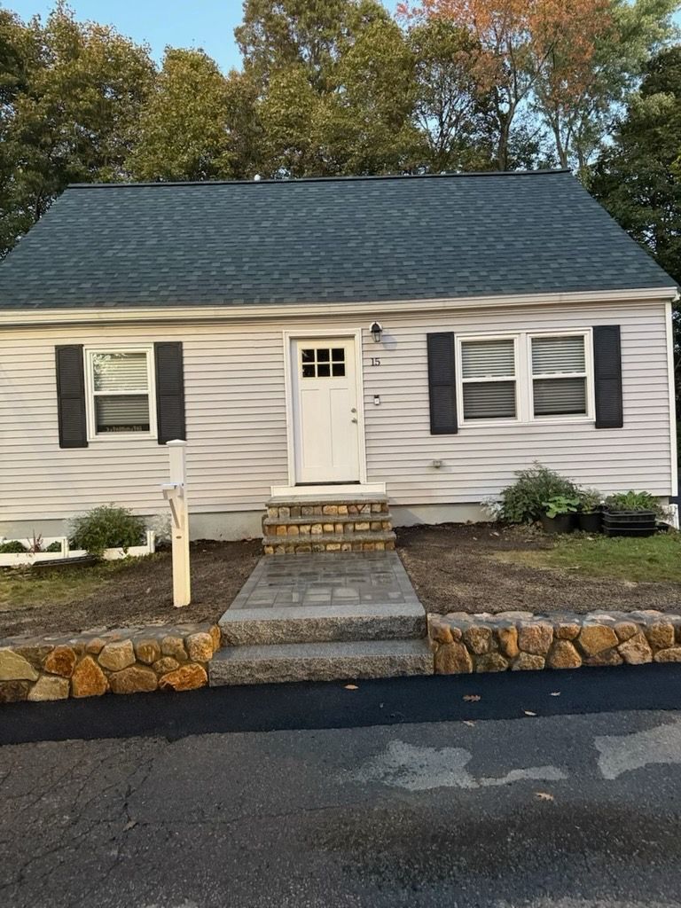 A white, single-story house with a green roof, dark shutters, and stone retaining walls along a paved driveway.