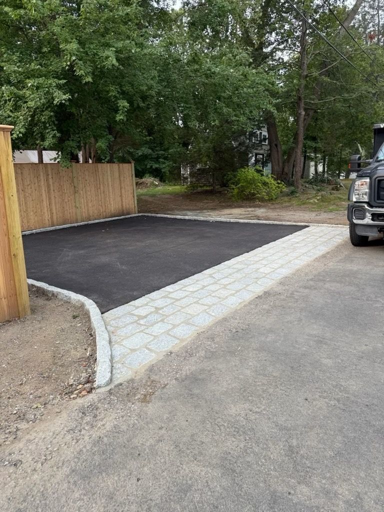 A newly paved asphalt parking pad bordered by light gray concrete pavers next to a wooden fence.