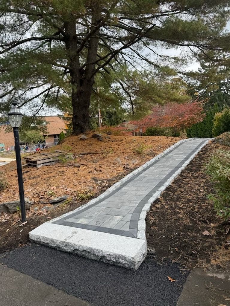 A paved stone walkway with granite border stones leads up a grassy slope toward a tree, next to a black lamppost.