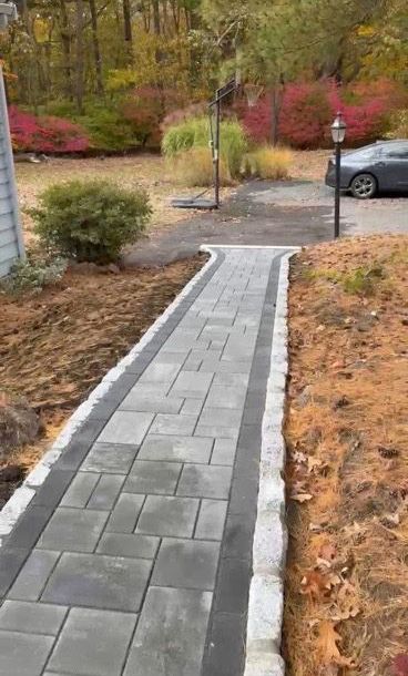 A new paved walkway with gray center pavers and a dark border, framed by light stone edging, leading to a driveway.