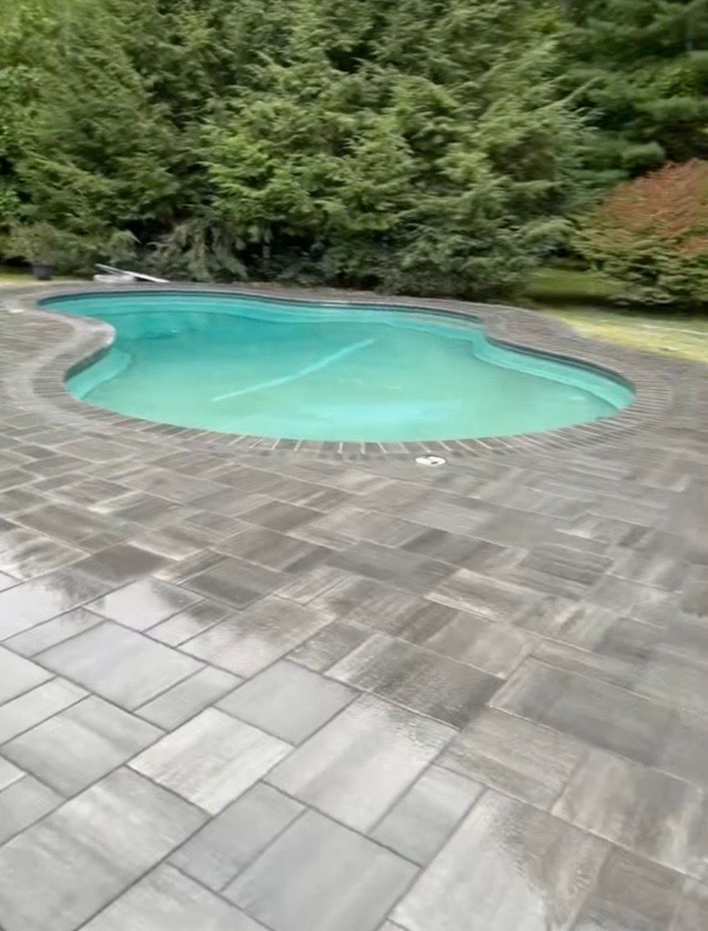A kidney-shaped swimming pool with turquoise water surrounded by a gray paver patio and lush green trees in the background.