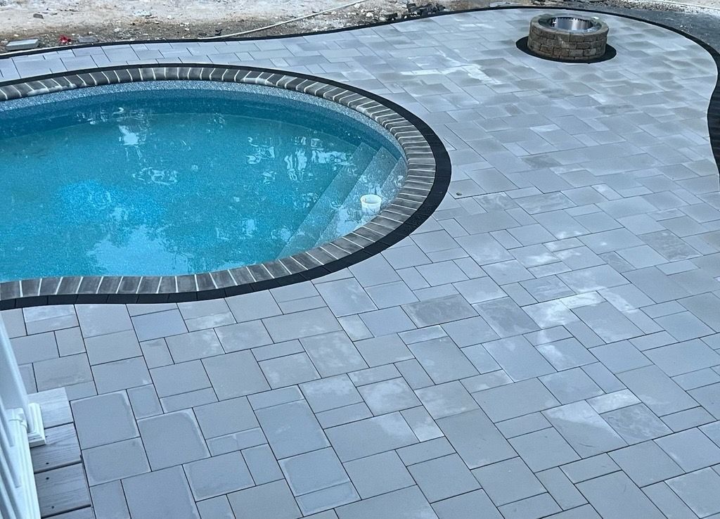A round swimming pool with dark stone coping sits in a gray paver patio featuring a stone fire pit.