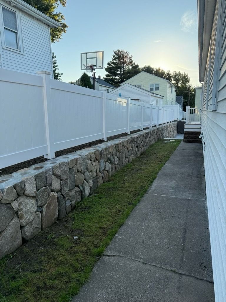 A stone retaining wall topped with a white vinyl fence runs along a narrow concrete path between two houses.