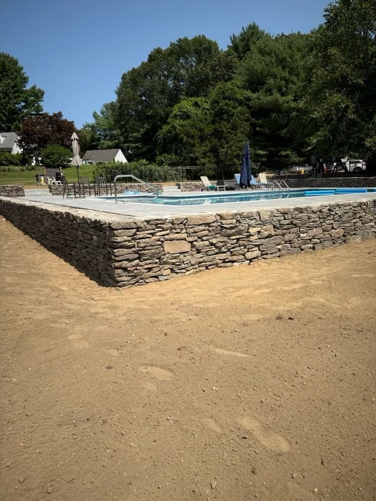 An elevated swimming pool framed by a textured stone wall, set in a yard with recently leveled sand.