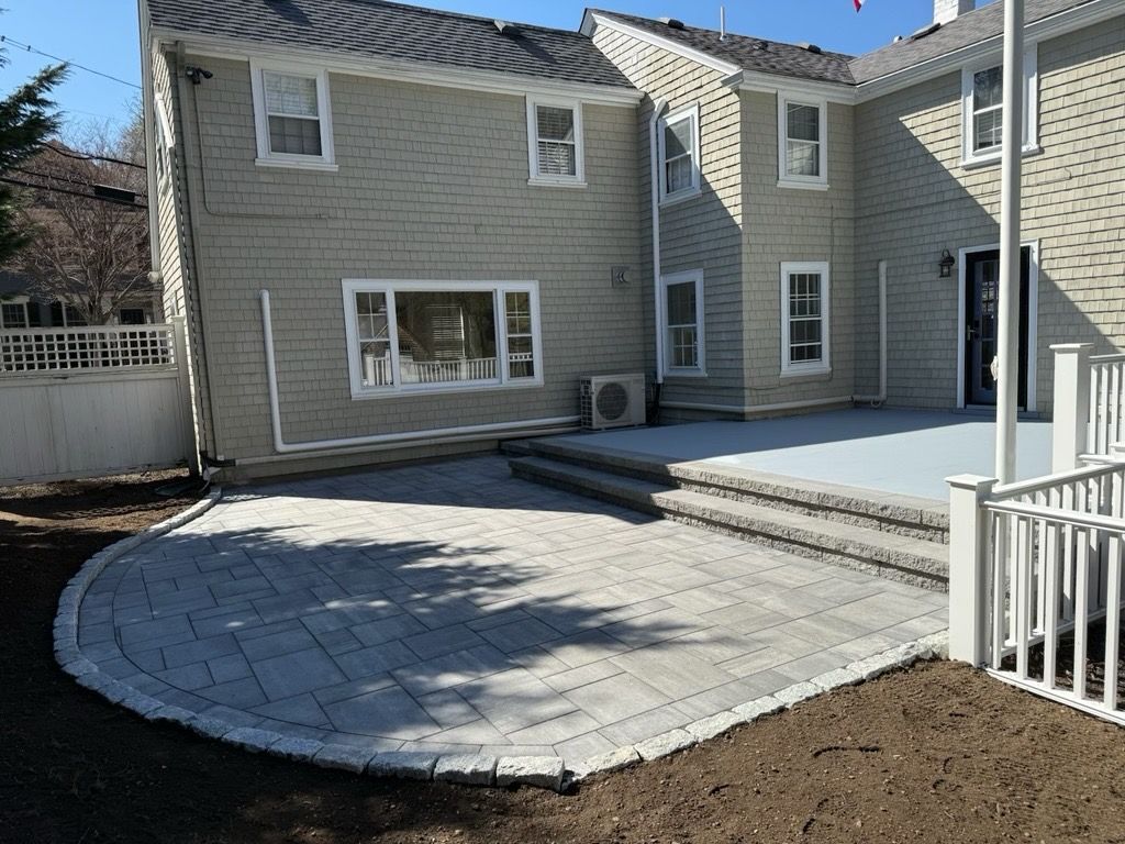 Backyard patio with curved paver area, steps to a deck, and a light-colored house.