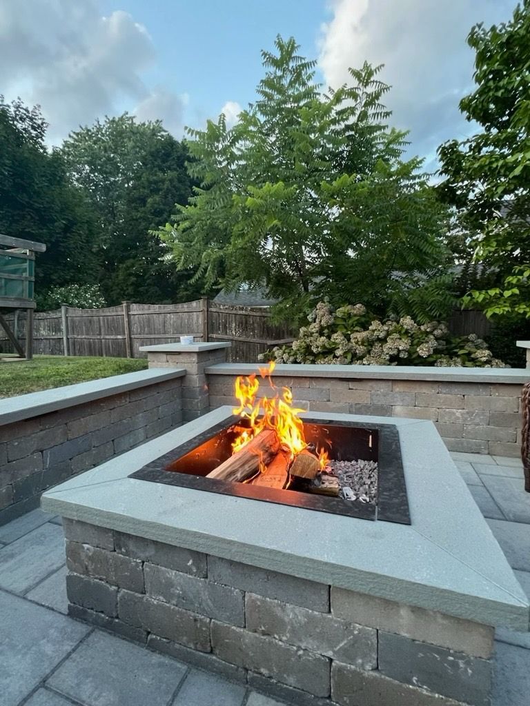 A fire pit with burning logs on a stone patio in a backyard setting.