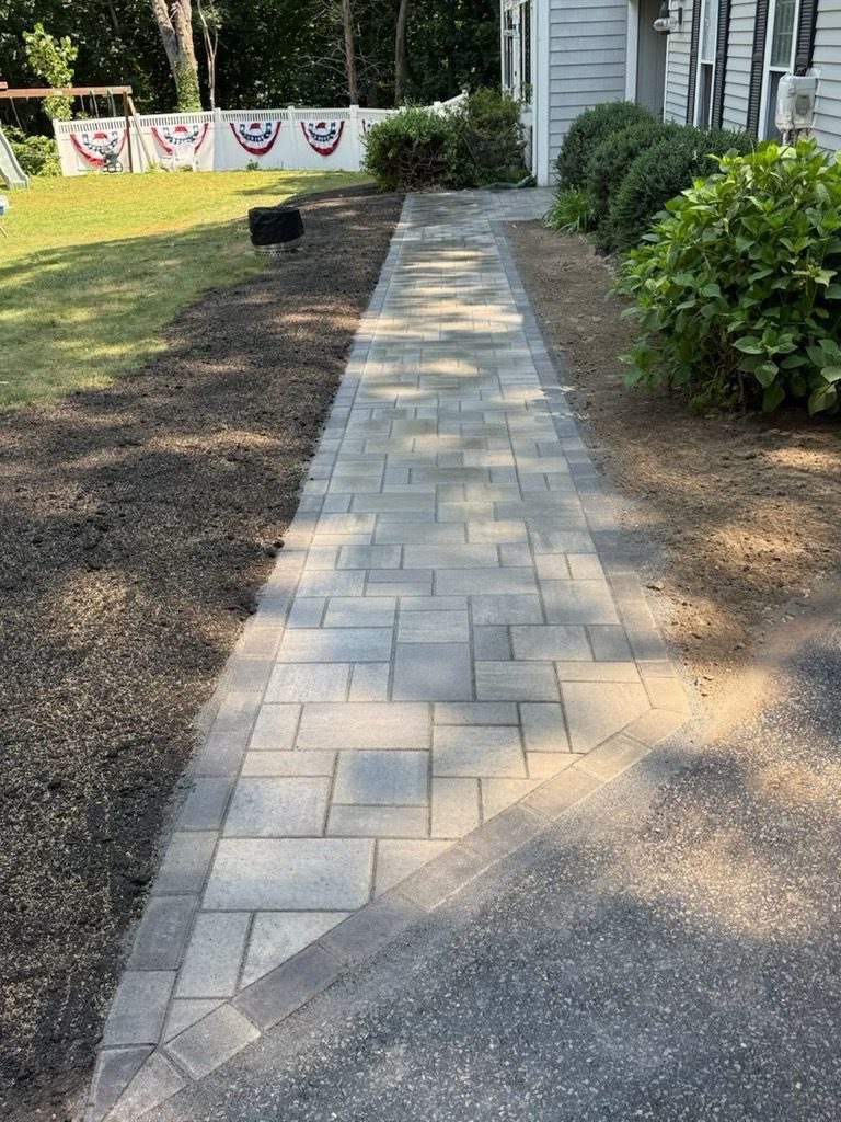 A paved walkway leads to a house entrance, with a bordering driveway and side garden.