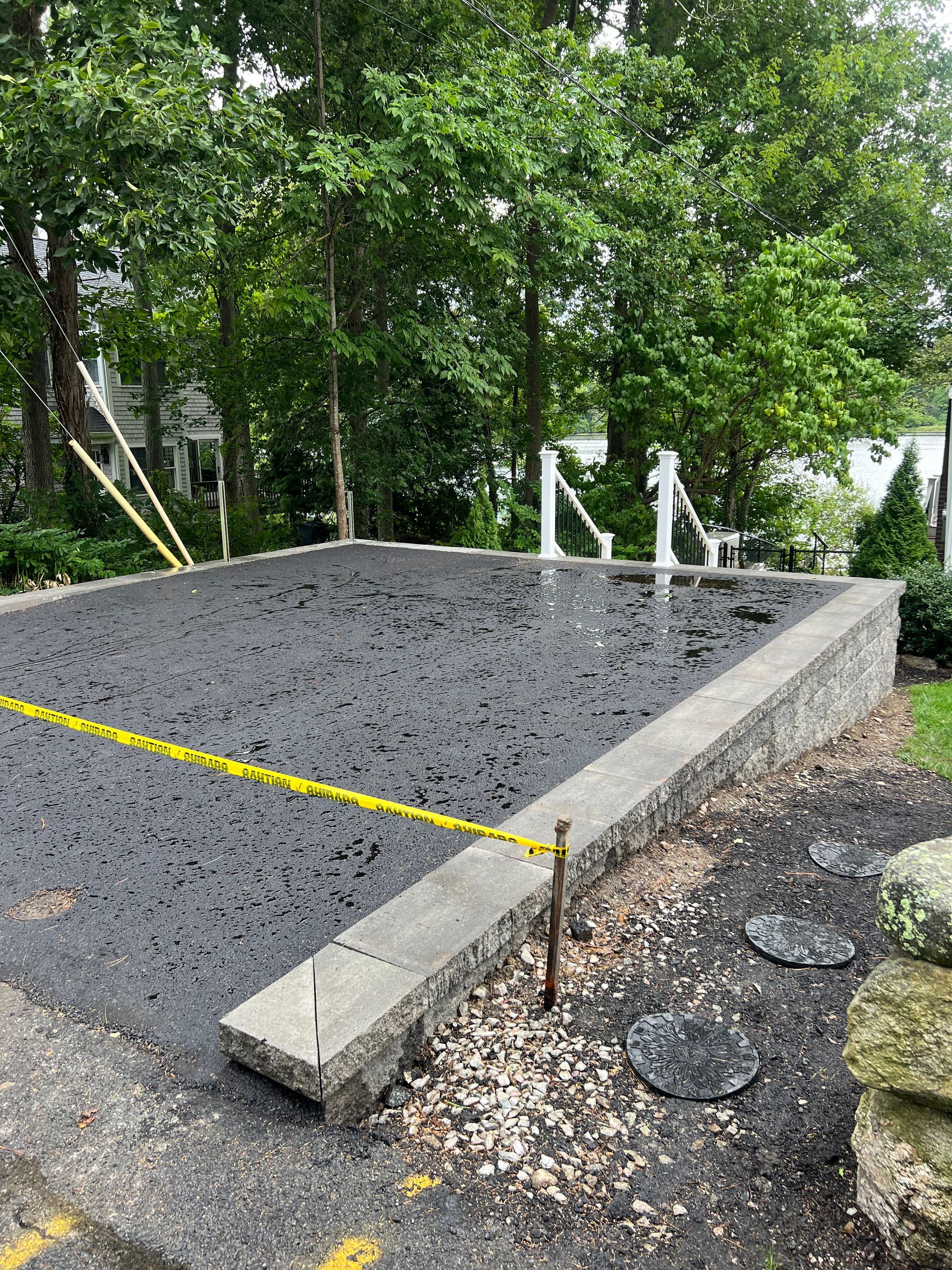 Asphalt-paved area with stone border and caution tape, surrounded by trees and a white fence.