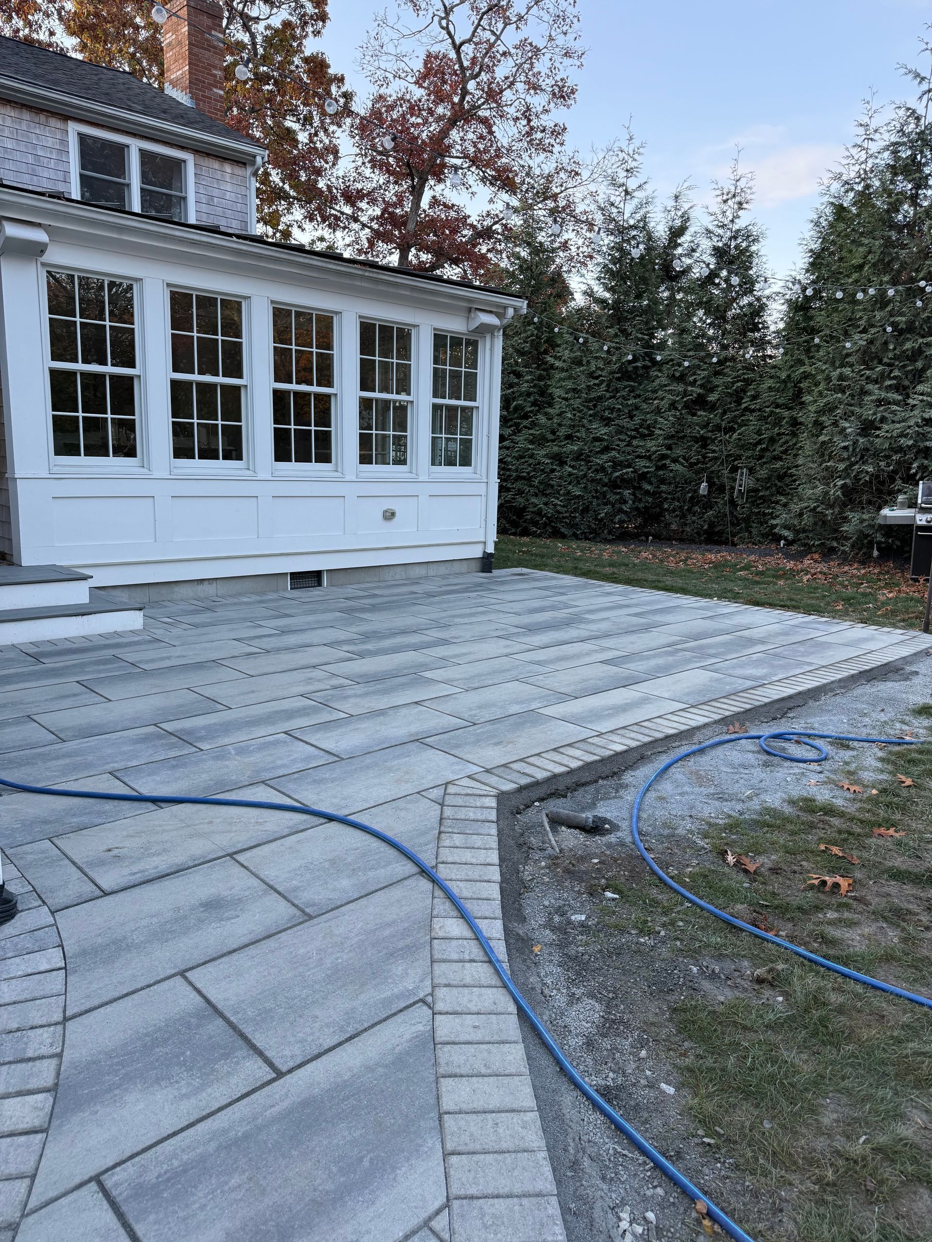 A gray stone patio with a blue hose extends from a white sunroom of a house.
