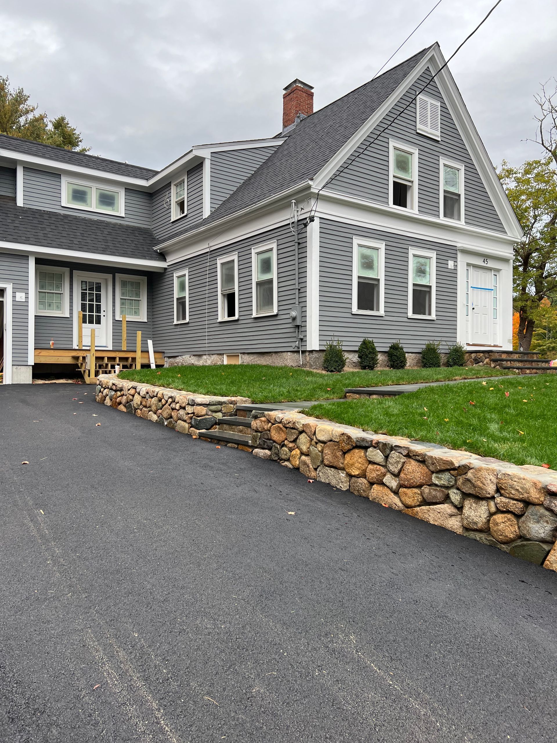 Gray house with asphalt driveway, stone wall, and green lawn.