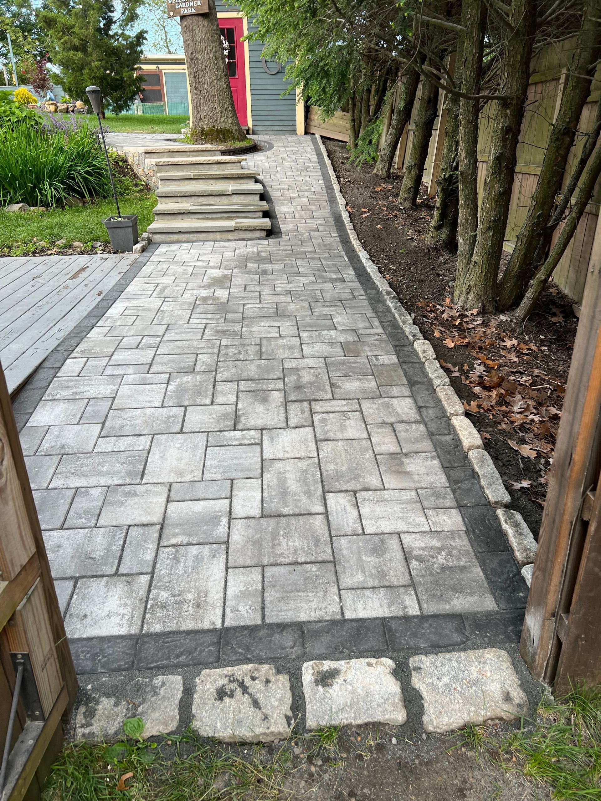 Stone path leading to steps and a garage, edged with black and lined with trees.