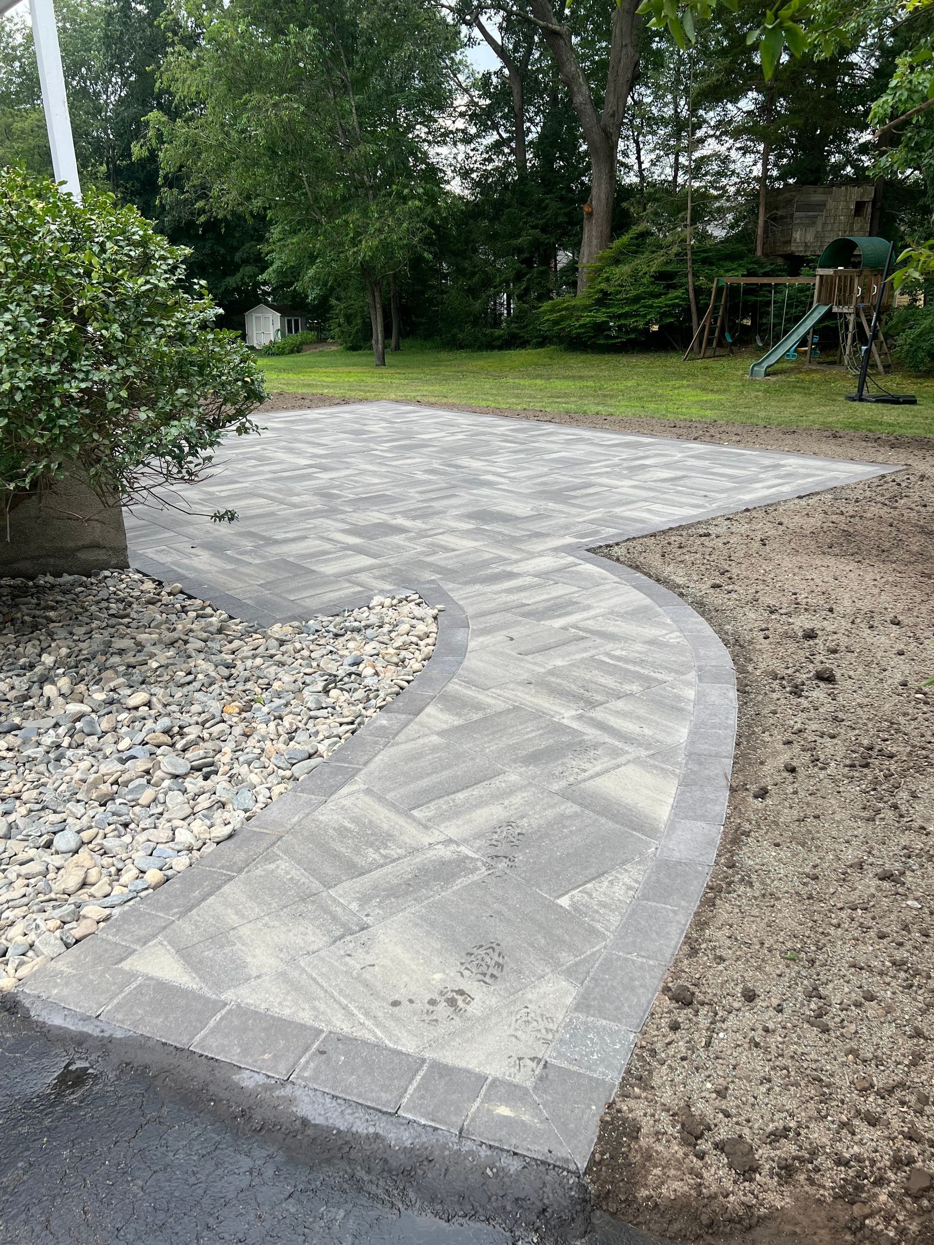 Stone patio and curved walkway in a yard; gray pavers, gravel border, grassy areas, trees.