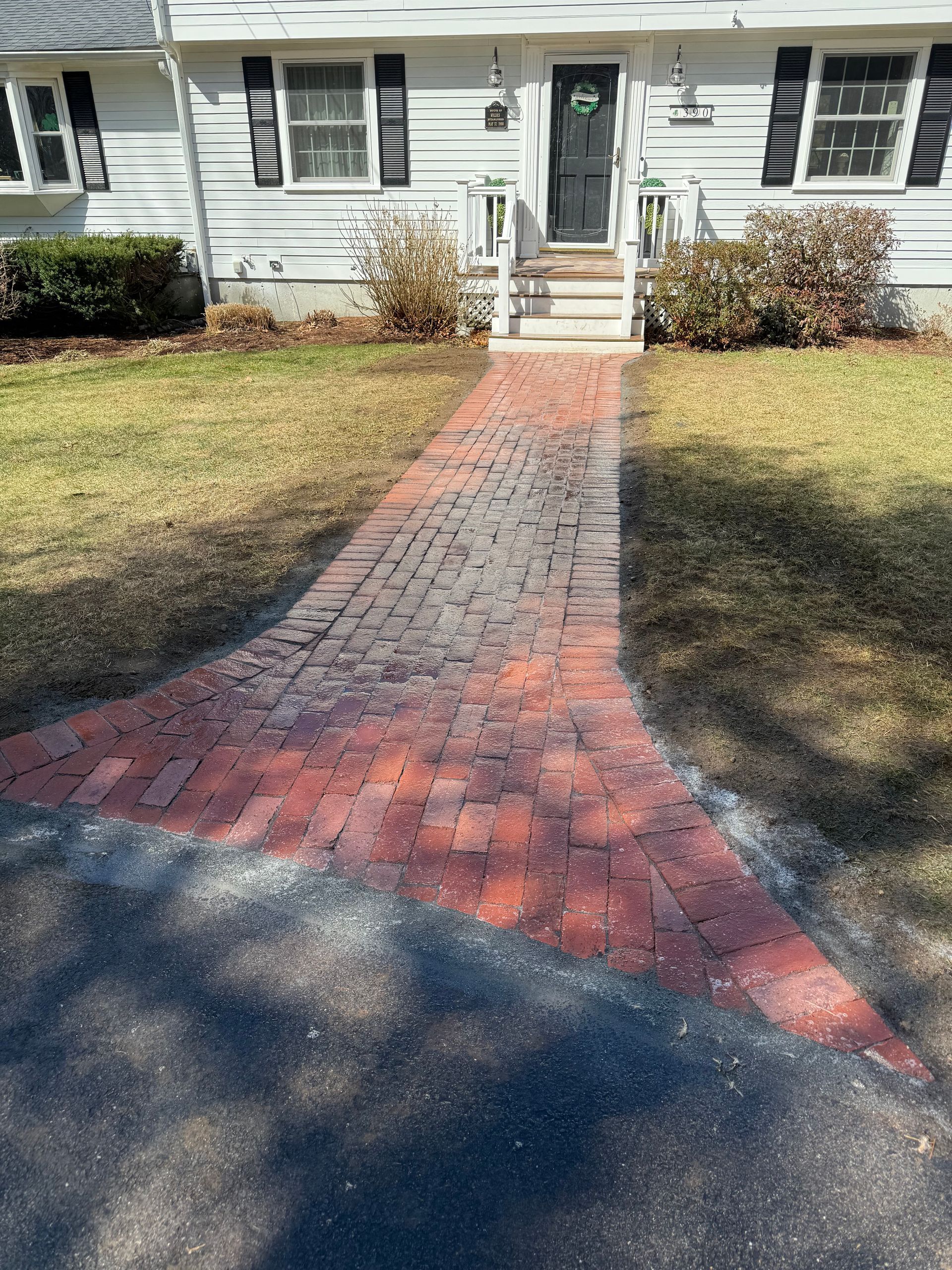 Brick path leading to a house with black door and bushes on either side.
