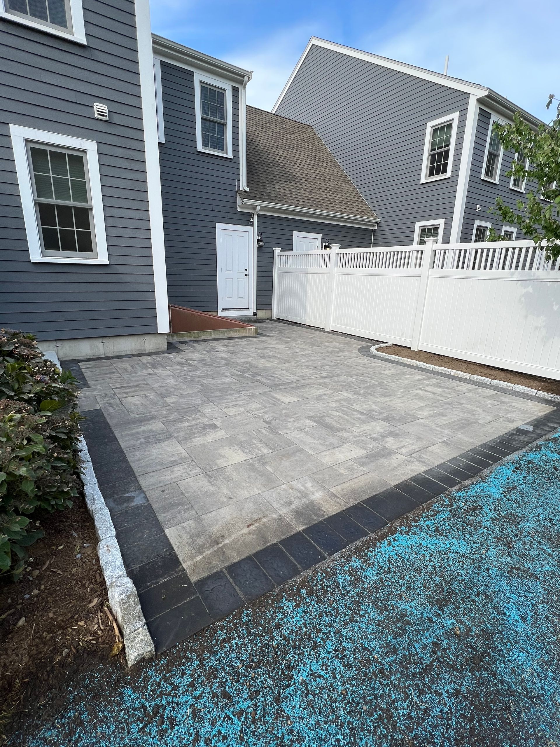 Paved patio with gray and black bricks next to a house with white fence; blue ground cover in front.