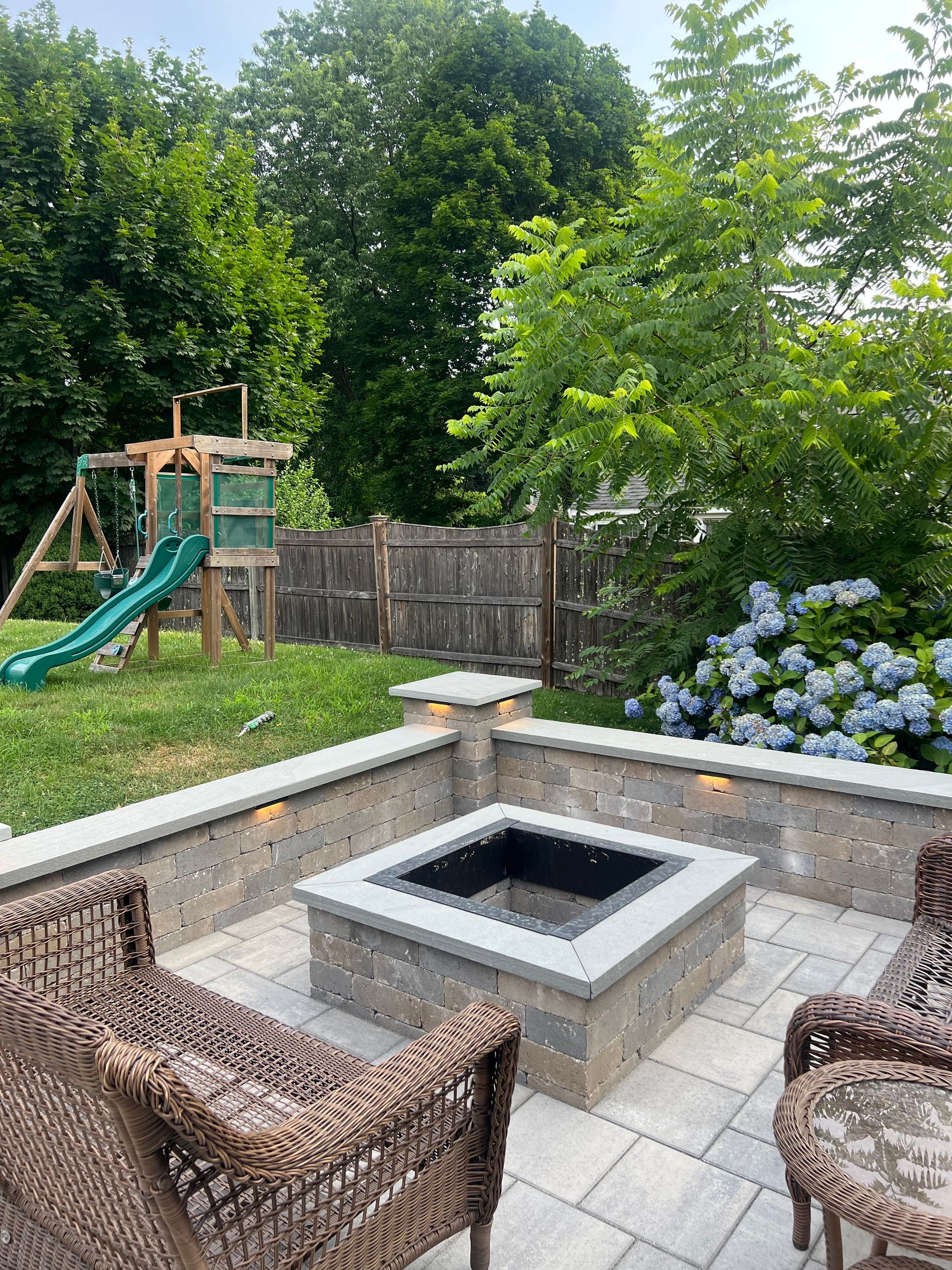 Backyard patio with fire pit, seating, playset, and blue hydrangeas.