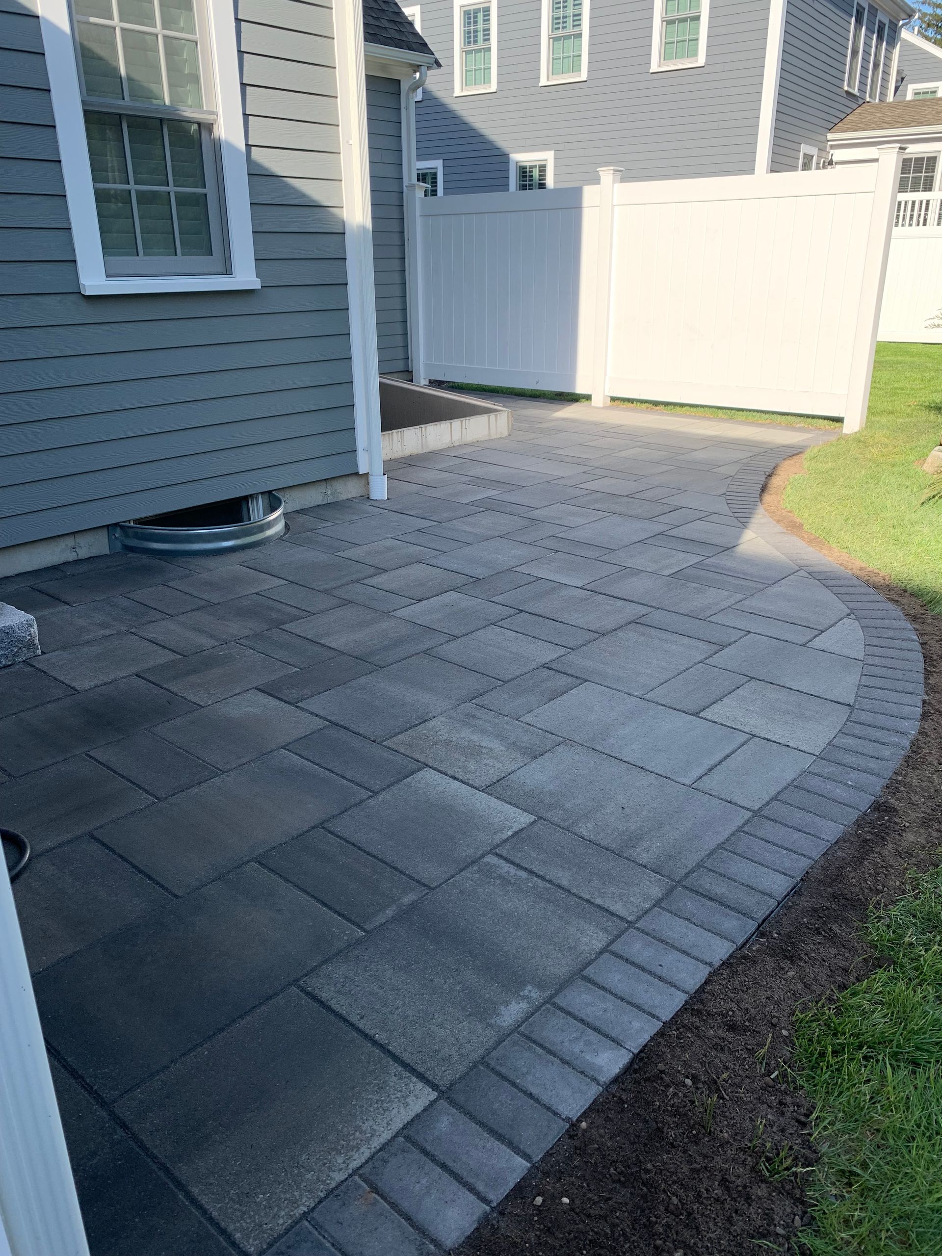 Paver patio with a brick border next to a house and white fence.