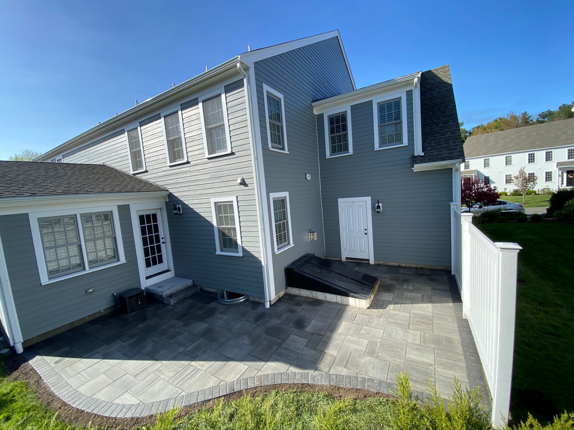 Back of a gray house with a patio, white fence, and blue sky.
