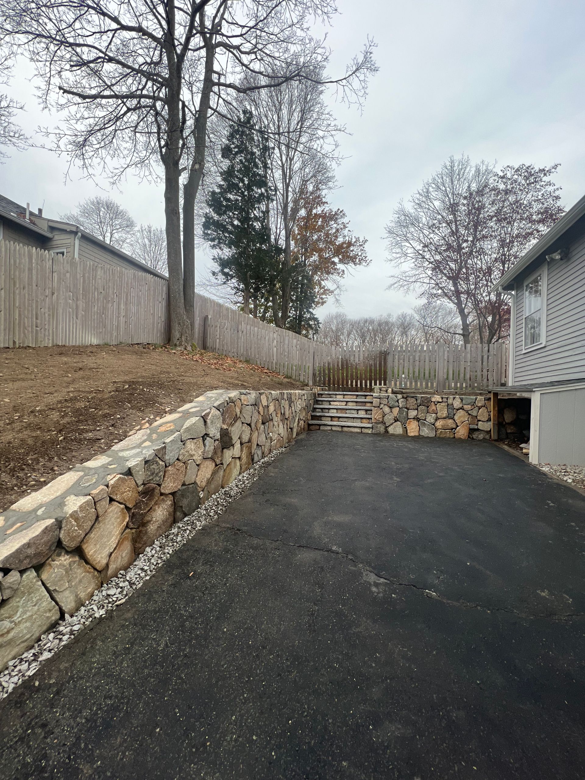 Driveway next to a stone retaining wall and fence. Trees in the background under an overcast sky.