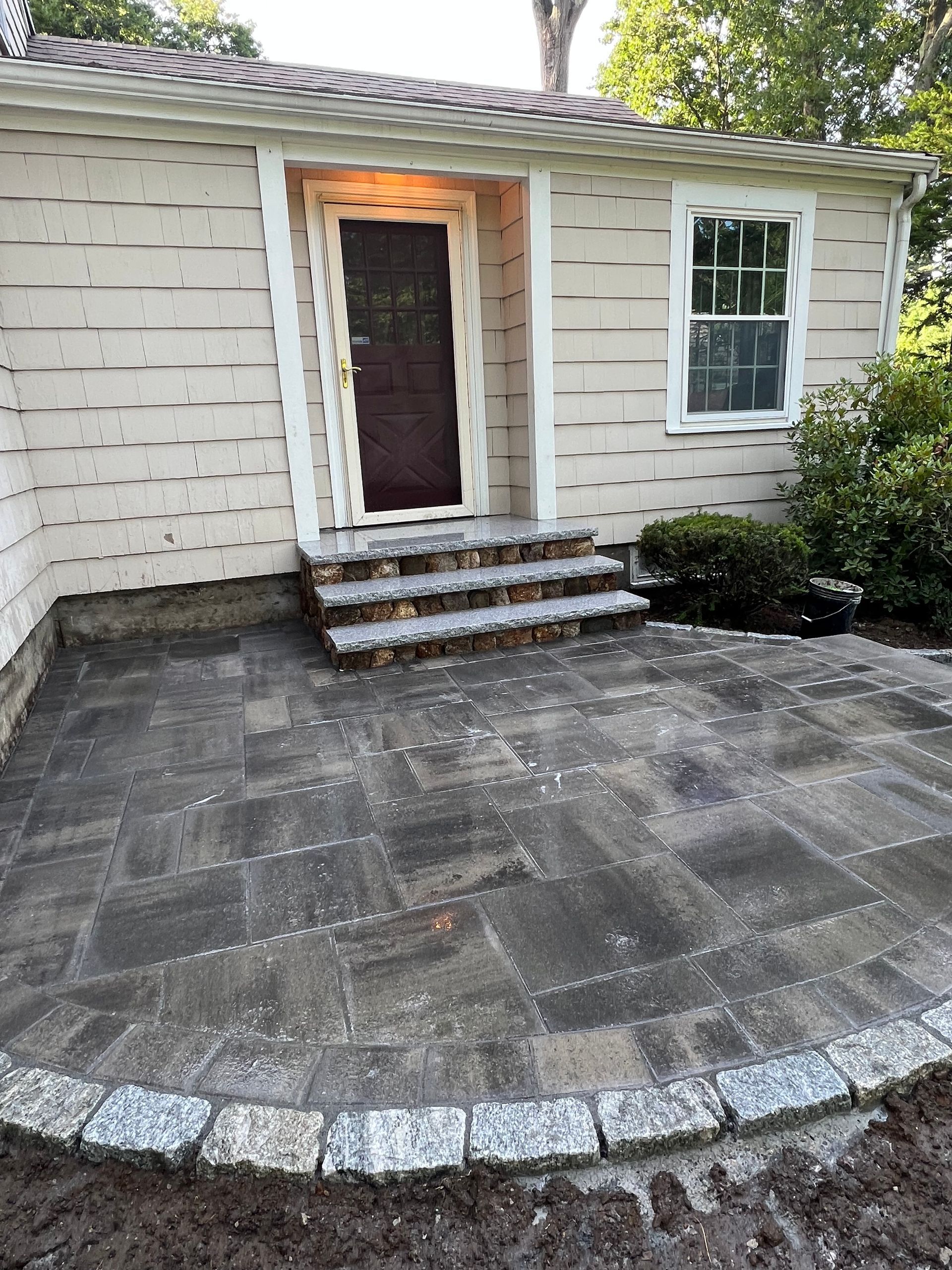 A newly paved patio with steps leading to a house entrance. Gray pavers with a granite border.
