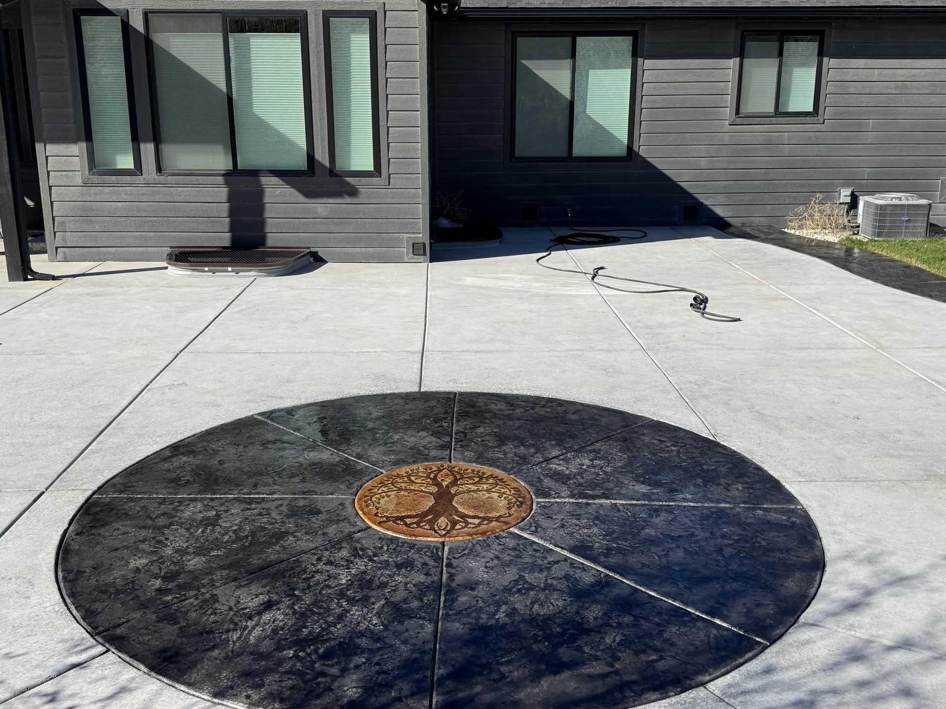 Concrete patio with a dark circular design and bronze emblem, in front of a gray house with windows.