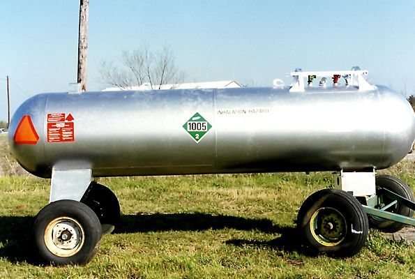 Silver propane tank on wheels in a grassy field, with hazard labels and a green hose attached