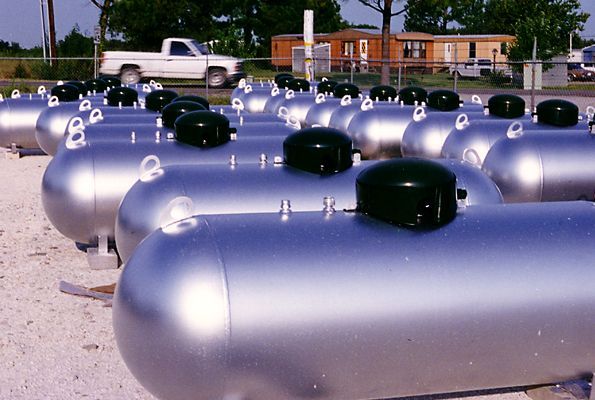 Rows of silver cylindrical tanks with black domes parked in an outdoor lot, with a pickup truck and building behind.