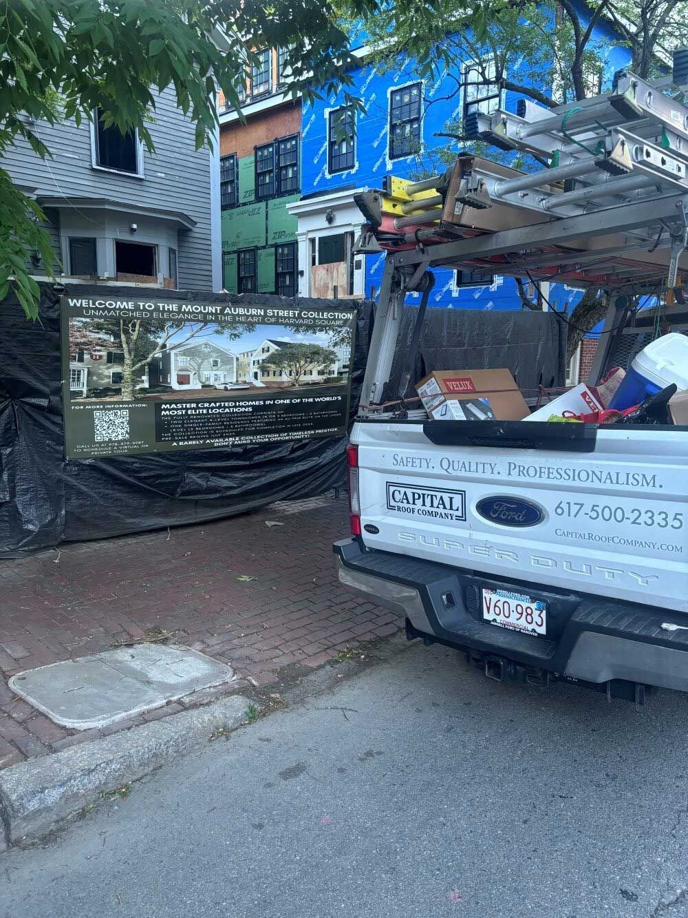 A work truck with a sign parked on a city street, houses with siding work in progress are behind it.