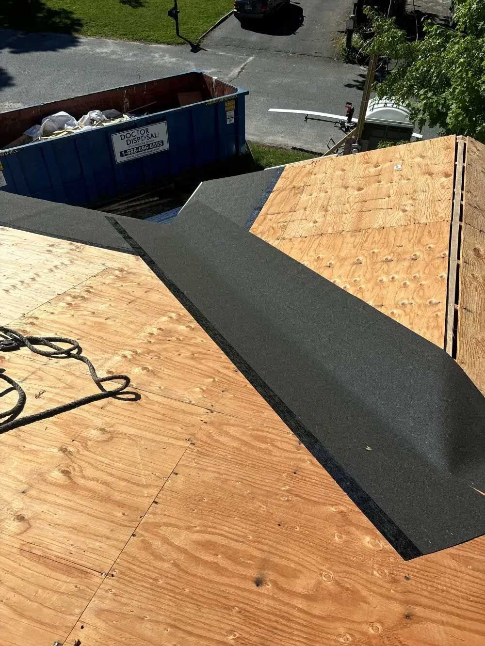 A new roof with black underlayment in place; plywood visible, blue dumpster in the background.