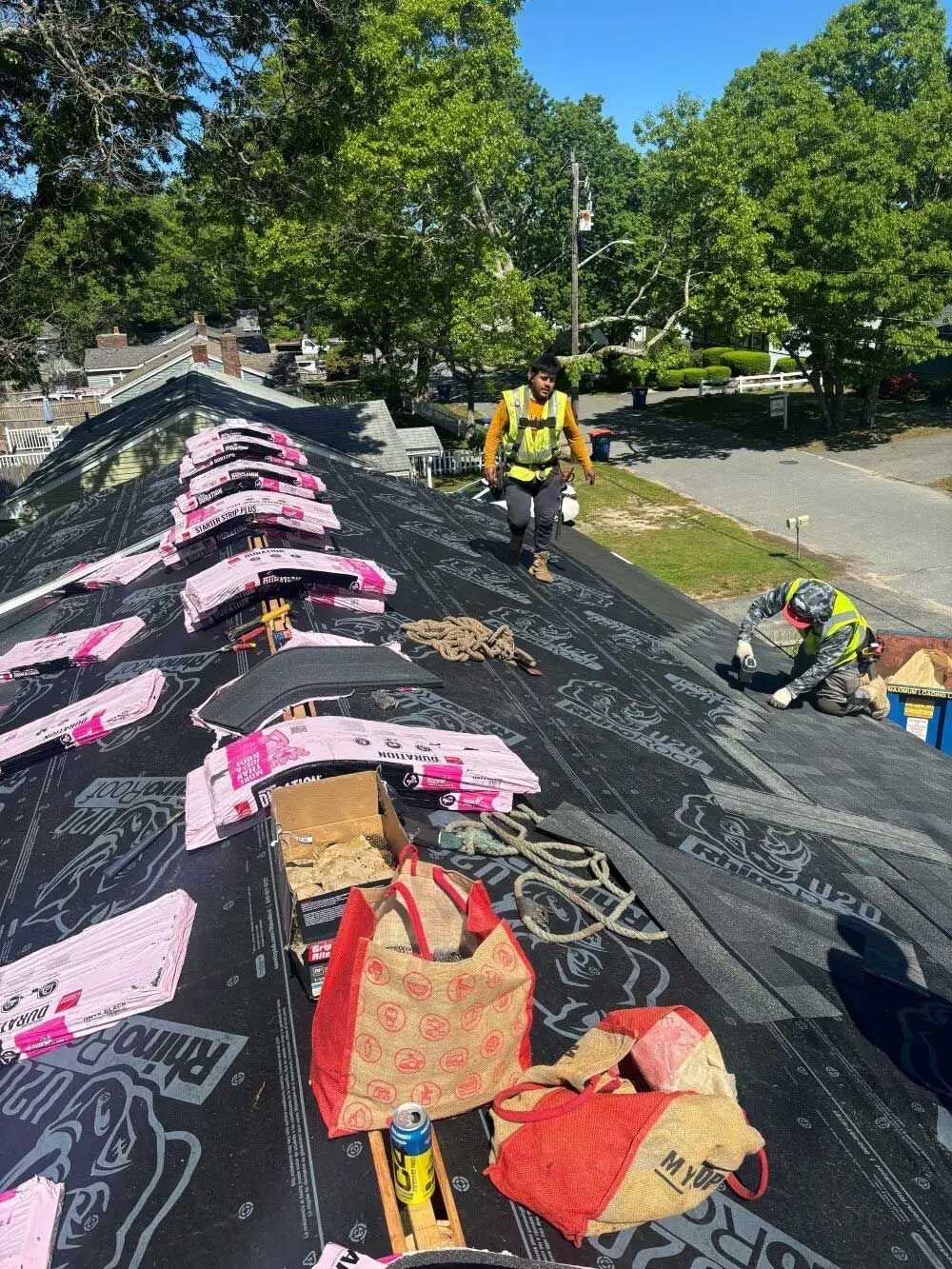 Roofers installing shingles on a roof, with tools, safety gear and bags visible. Sunny day.