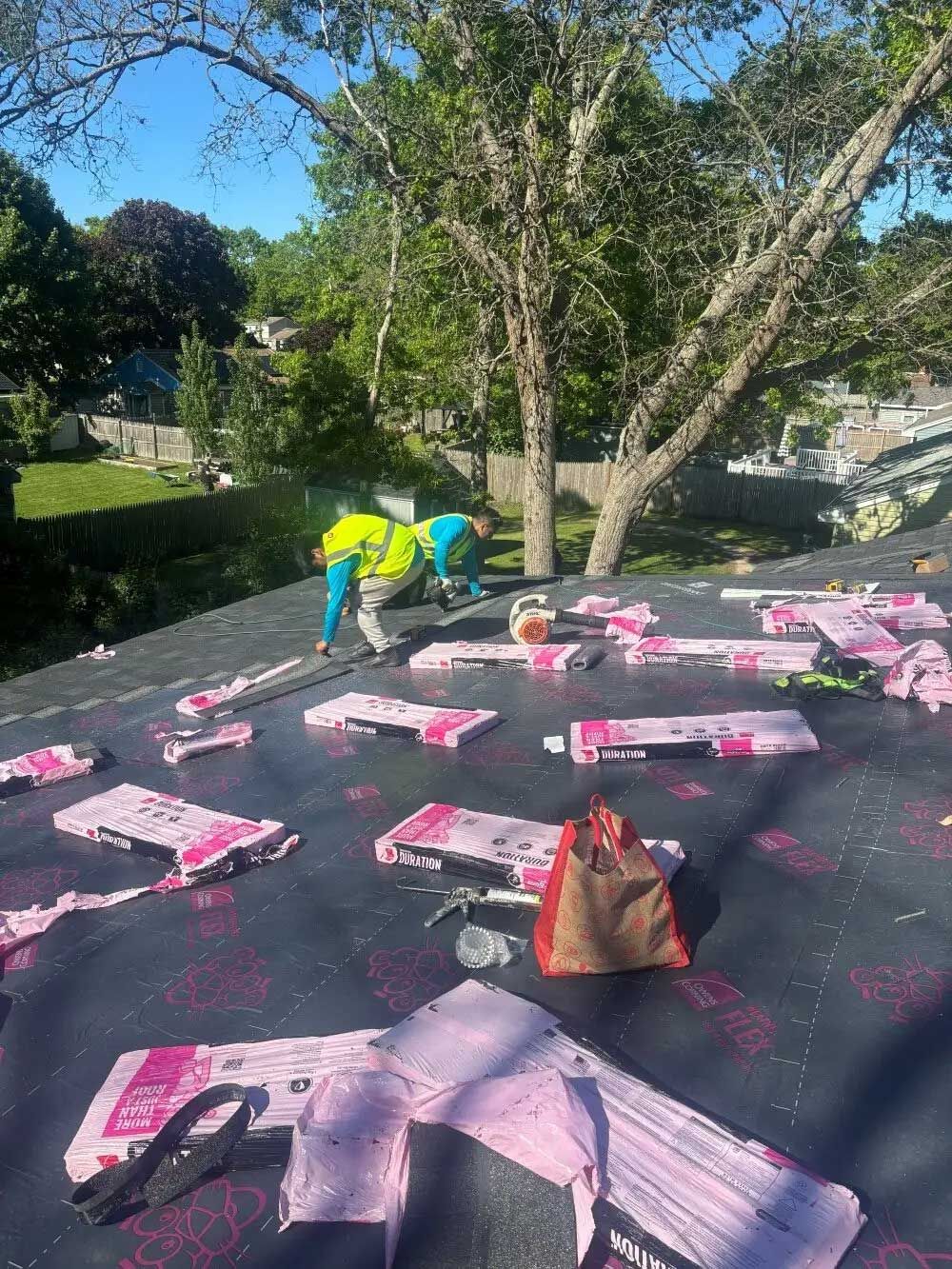 Roofers installing pink insulation on a roof under a sunny sky, trees and houses in the background.