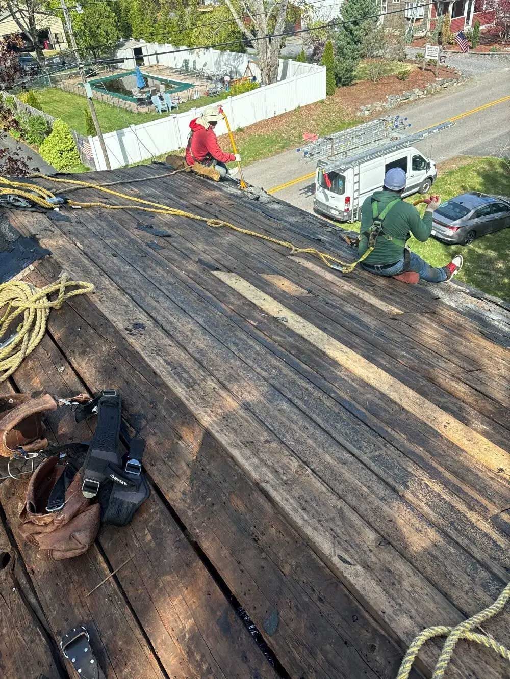 Roofers on a weathered rooftop, using safety harnesses. One hammers, the other rests; a van and road are visible.