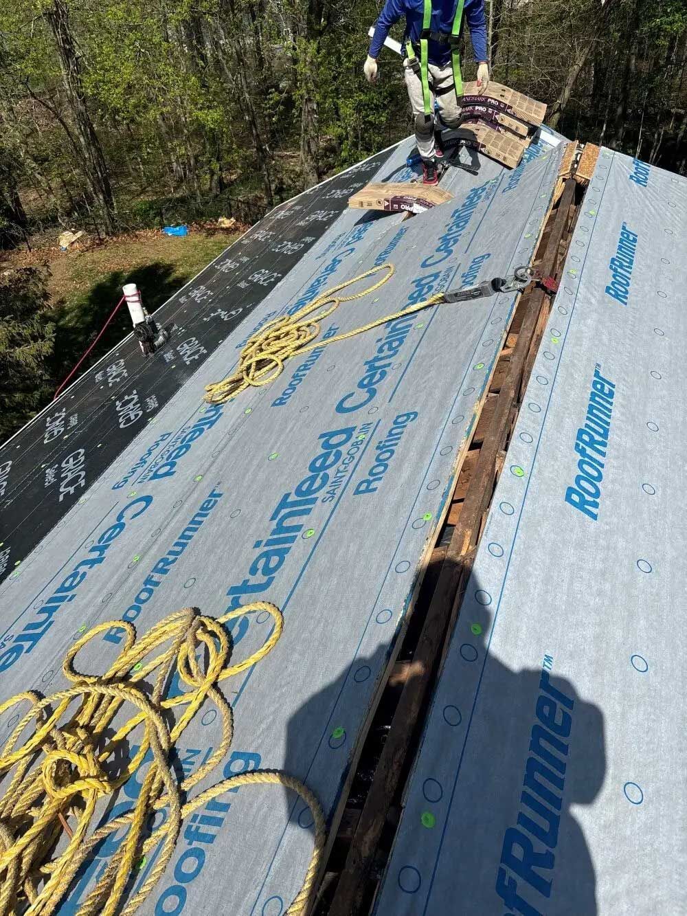 Roofer on a roof wearing safety gear, laying roofing material. Blue underlayment, yellow rope, sunny outdoors.