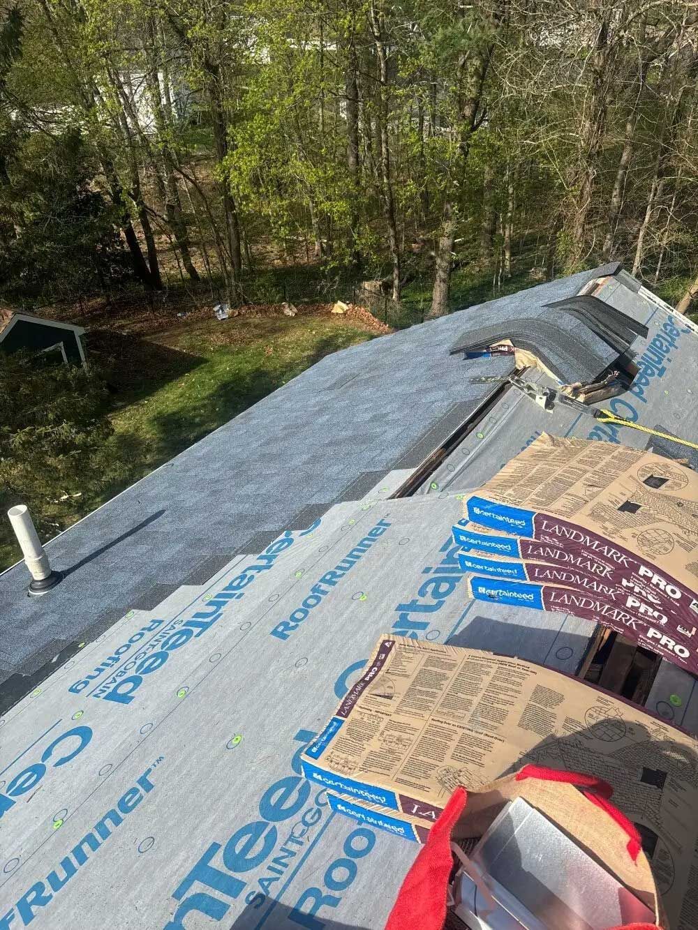 Roof being worked on, with new underlayment and asphalt shingles. Trees and grass in background.
