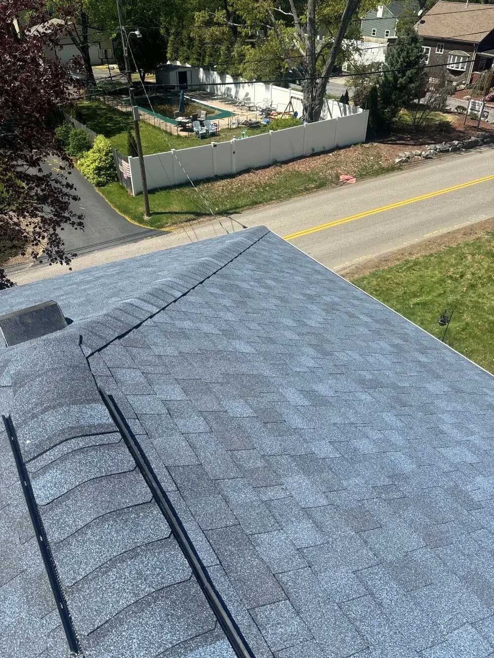 View of a blue shingled roof with a street and yard visible below on a sunny day.