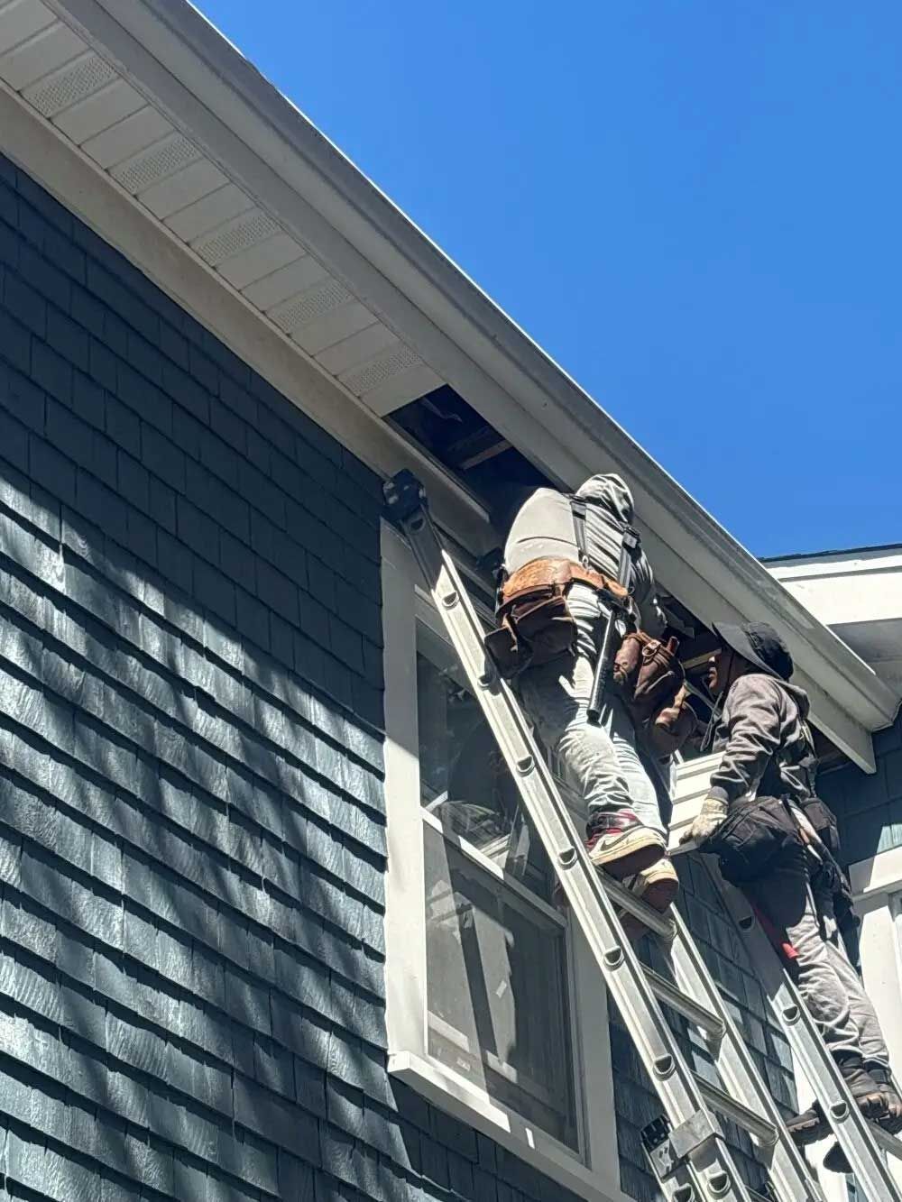 Two workers on a ladder repairing a house's soffit and fascia against a blue sky.