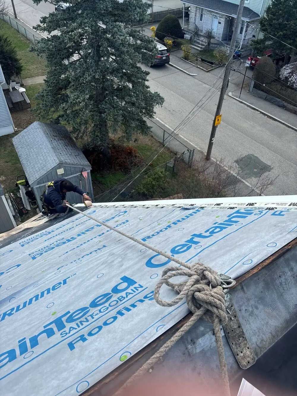 Roofer on a roof, secured by rope, working near a street with houses and a tree.