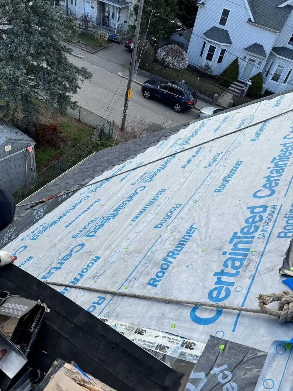 Roofer installing shingles on a house roof with safety rope, a street and other houses in the background.
