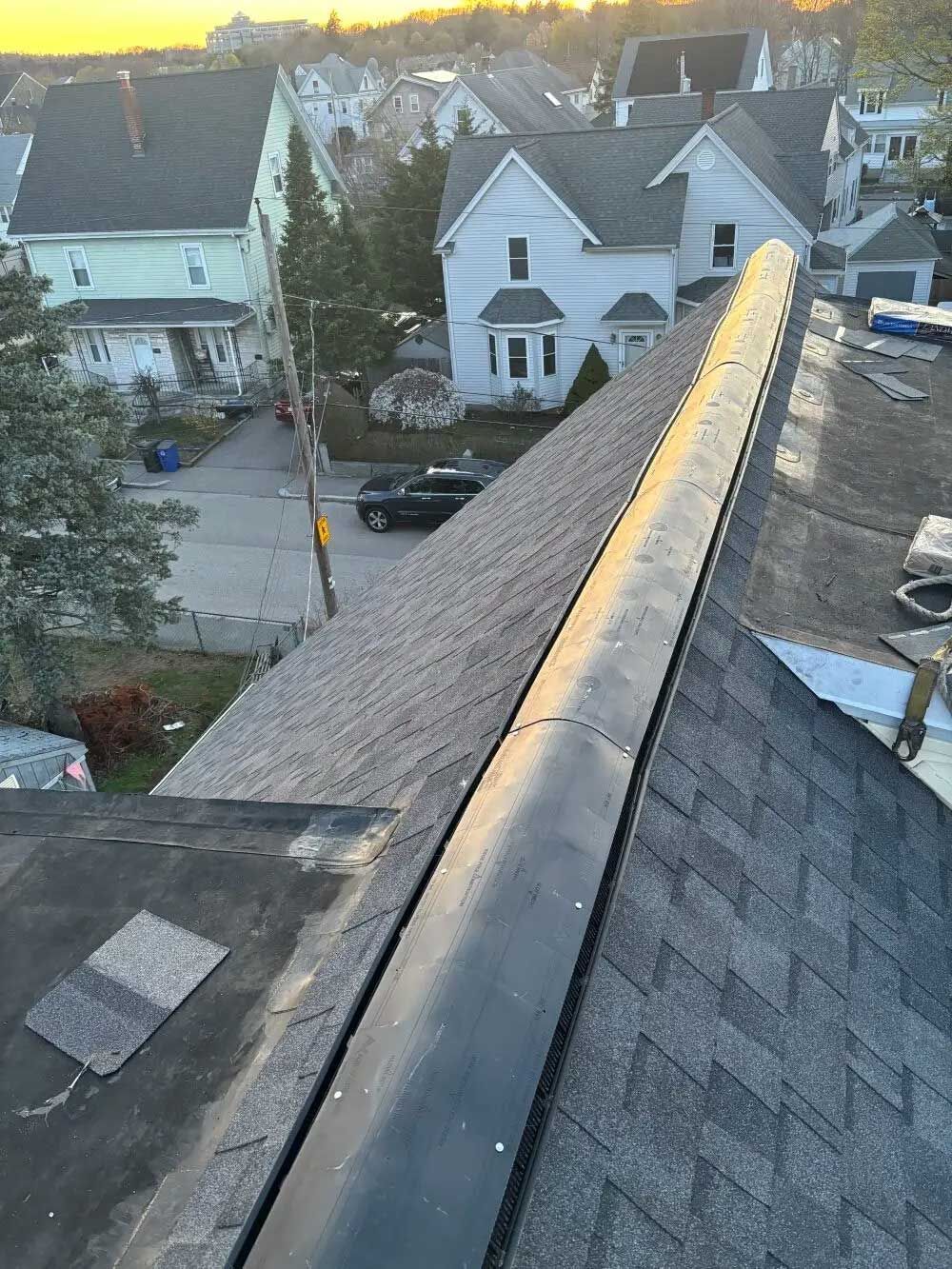 View of a rooftop with a black ridge cap, overlooking a neighborhood at sunset.