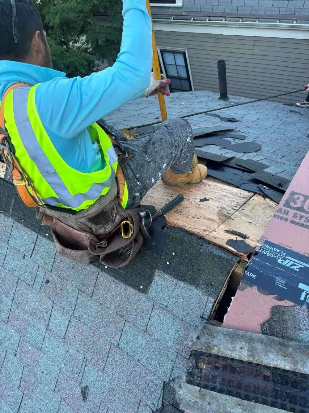 Roofer on a roof, wearing safety gear, holding a level, working on replacing shingles.