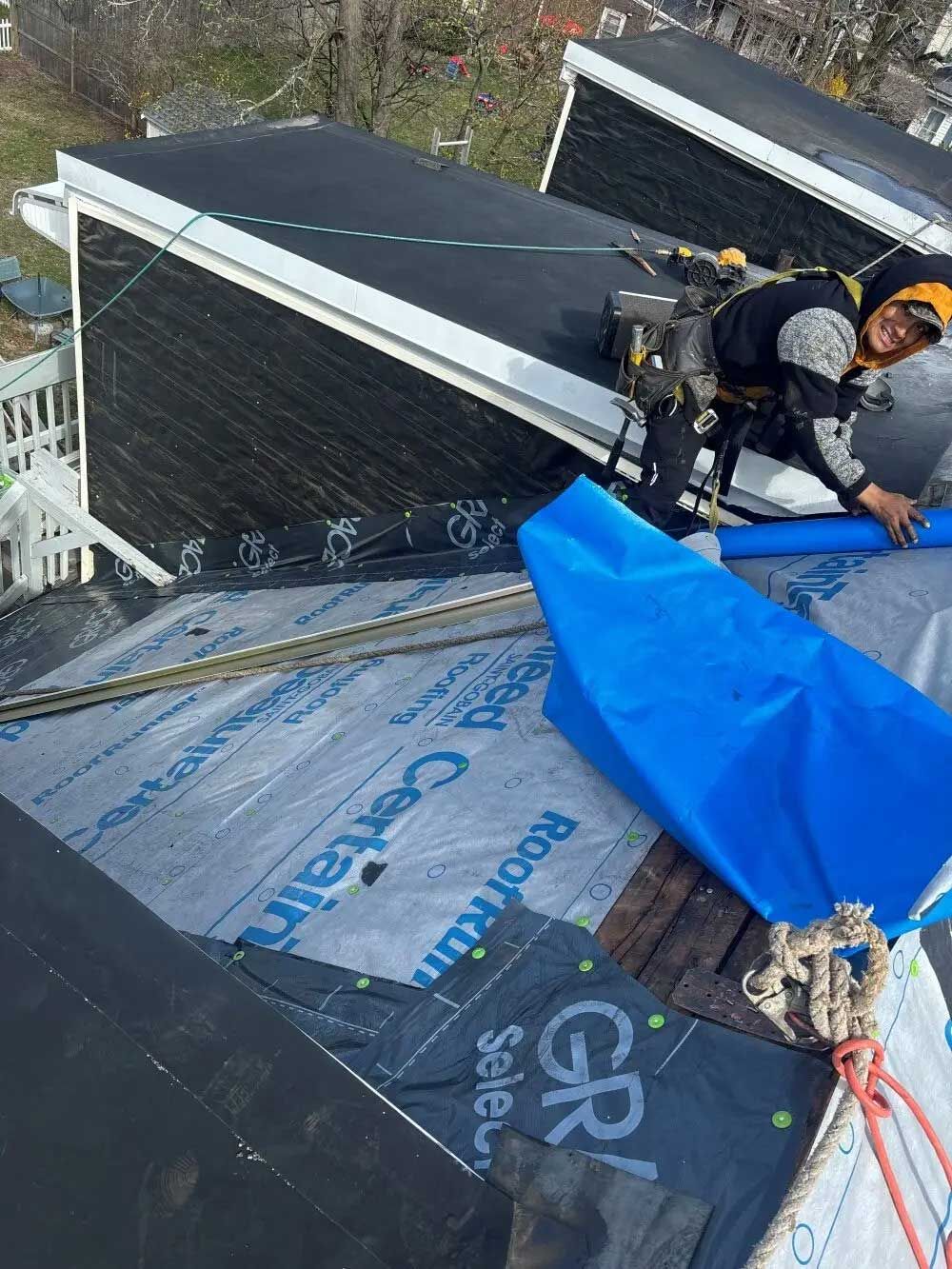 Roofer on a roof, smiling. Wearing safety gear, applying material, near a blue cover.