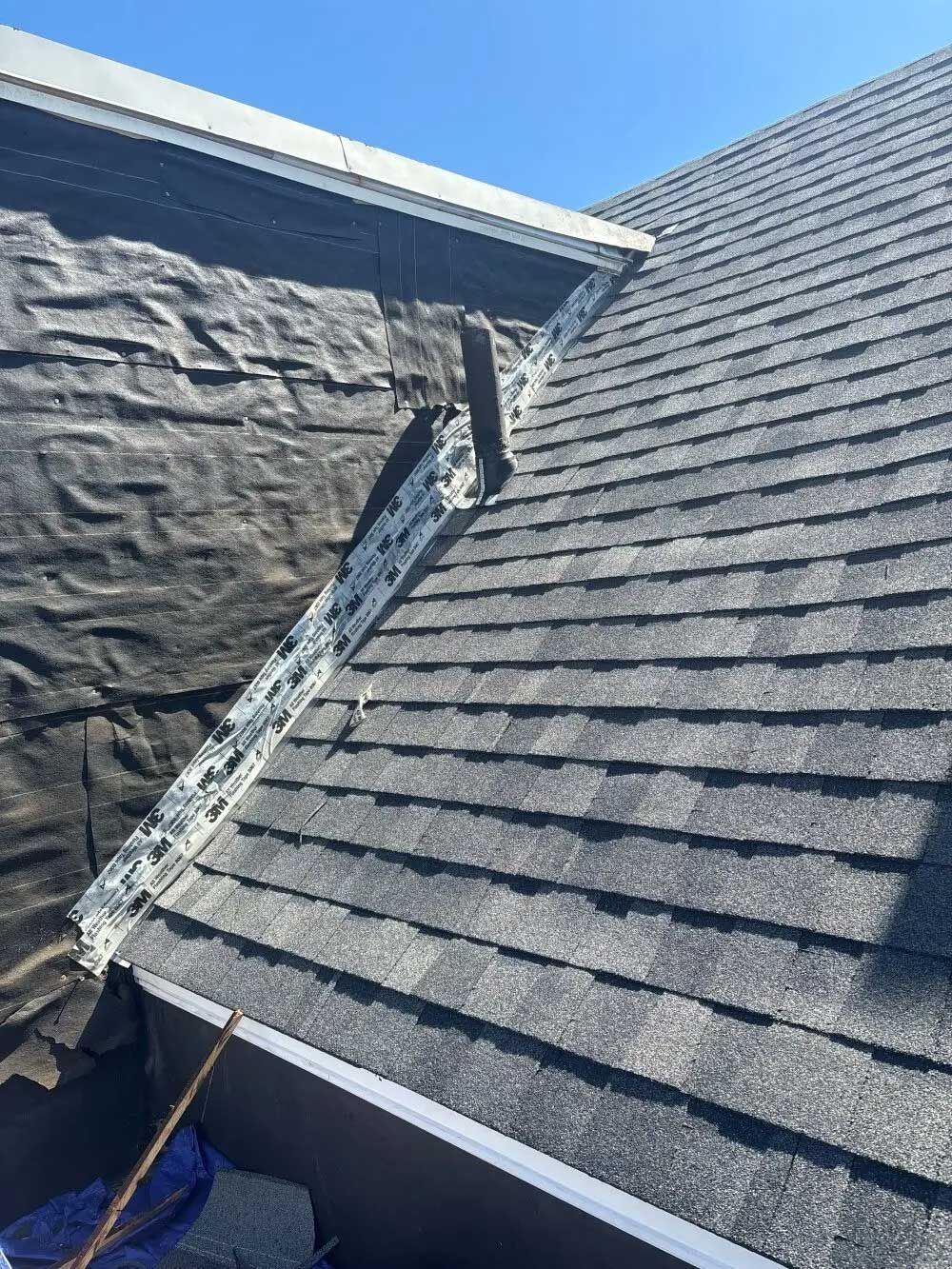 Roof with black shingles and vent pipe against a blue sky.